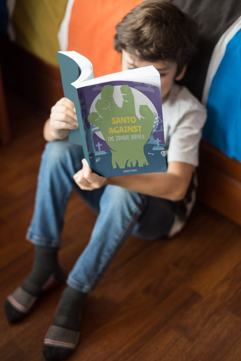 Boy Reading A Paperback Book Mockup Sitting Against His Bed