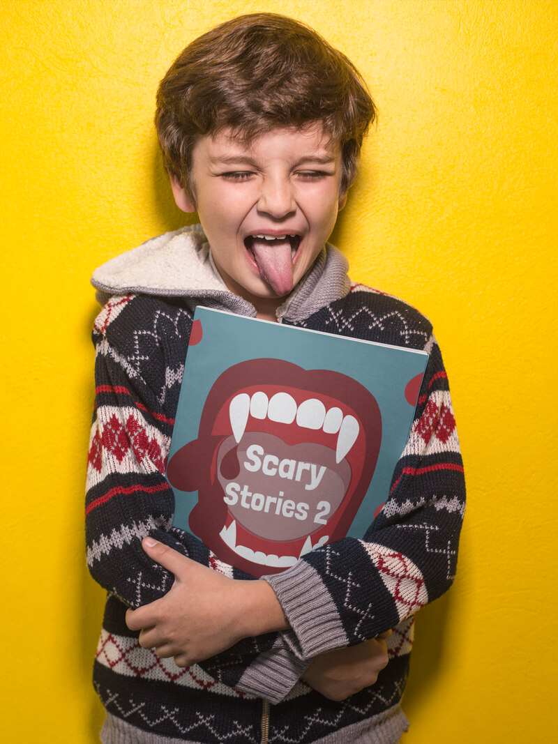 Boy Sticking His Tongue Out Holding A Book Mockup Against A Yellow Wall