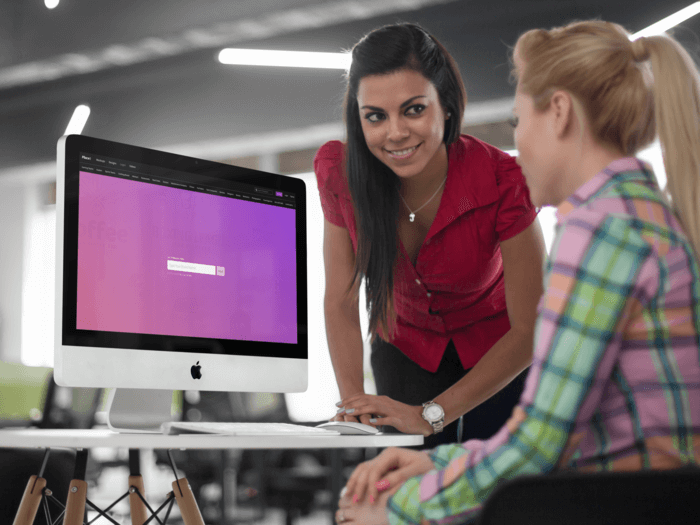 Two Young Businesswoman Chatting In Front Of An Imac Mockup Logo