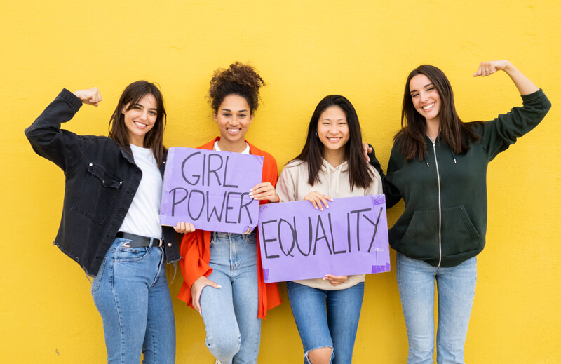 Group Of Multiethnic Young Women With Feminist Signs On A Yellow Background.