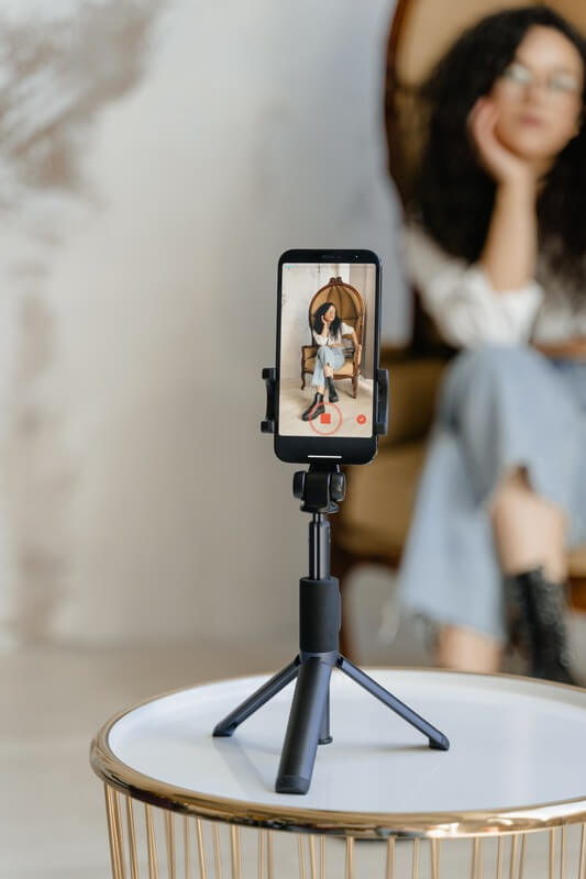 A Close Up Of A Phone Displaying The Tiktok App, With A Woman Blurred In The Background As She Records A Tiktok Video.