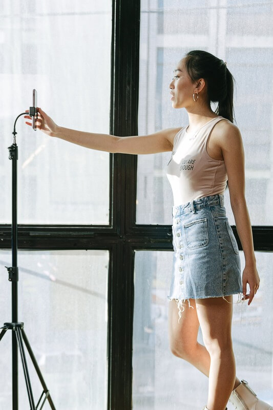 A Young Woman Standing In Front Of Her Phone, Learning How To Edit Tiktok Videos.