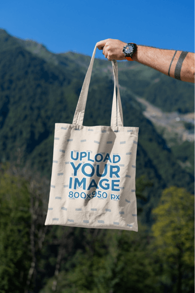 Mockup Featuring A Man's Hand Holding A Tote Bag Against A Natural Scenery