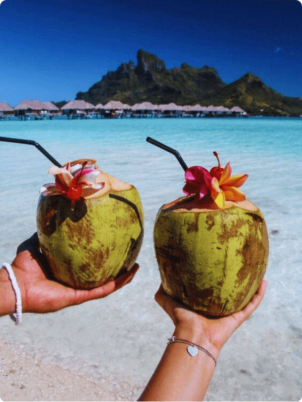 Two Coconut Beverages Held With A White Beach In The Background