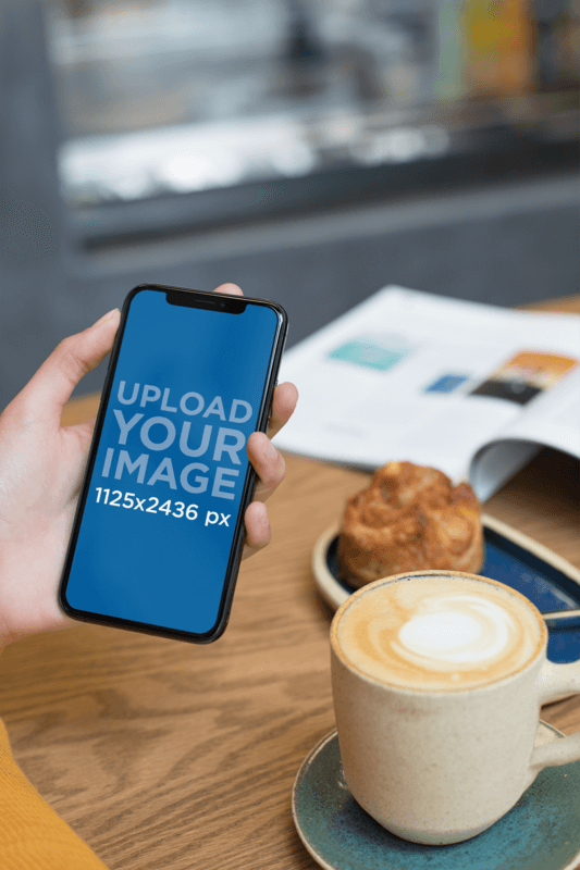 Mockup Of A Woman Holding An iPhone Xs While At A Cafe